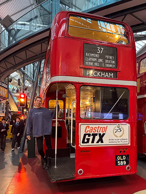 Наталя стоїть на задній платформі класичного лондонського даблдеккера типу RT. Одна з наймасовіших моделей автобусів. На листівці є така модель автобуса у святковій золотій лівреї на честь коронування Єлизавети ІІ у 1953 році — RT Gold. Докладніше про конкретний даблдеккер RT, що на фото: [www.ltmuseum.co.uk](https://www.ltmuseum.co.uk/collections/collections-online/vehicles/item/1981-520)